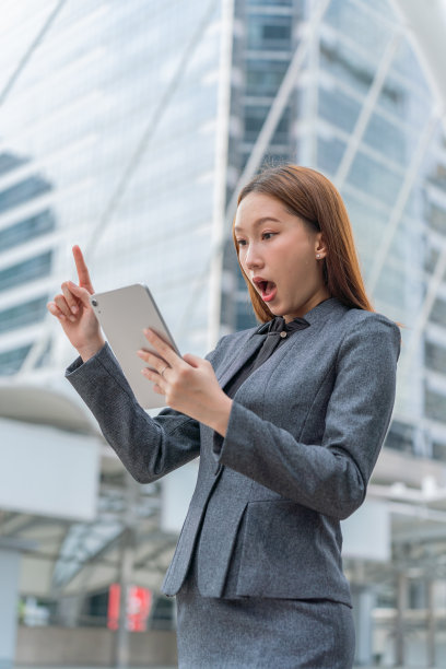一名年轻的亚洲女商人穿着灰色的办公套装，在商业区使用她的平板电脑时微笑着图片下载