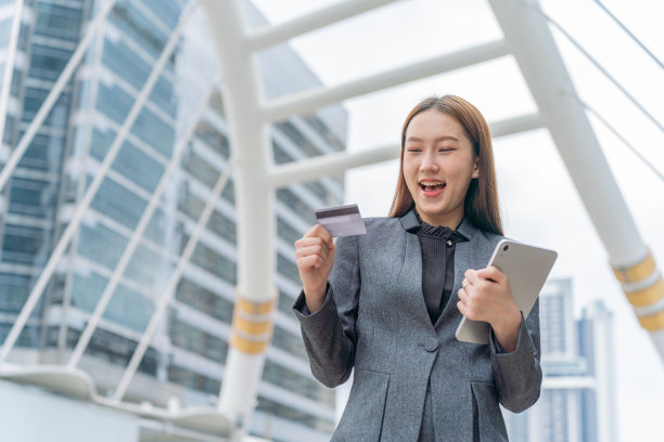 在一个商业区，一名身穿灰色西装的年轻亚洲女商人在使用她的信用卡通过她的数字平板电脑进行在线交易时自信地微笑着图片下载