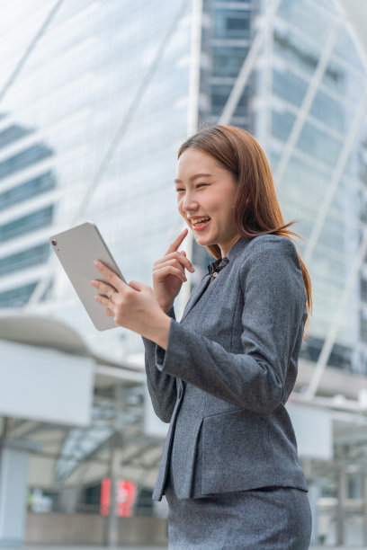 一名年轻的亚洲女商人穿着灰色的办公套装，在商业区使用她的平板电脑时微笑着图片下载