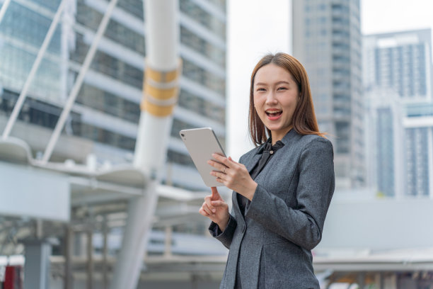 一名年轻的亚洲女商人穿着灰色的办公套装，在商业区使用她的平板电脑时微笑着图片下载