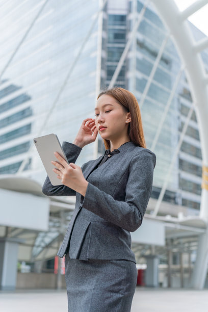一名年轻的亚洲女商人穿着灰色的办公套装，在商业区使用她的平板电脑时微笑着图片下载