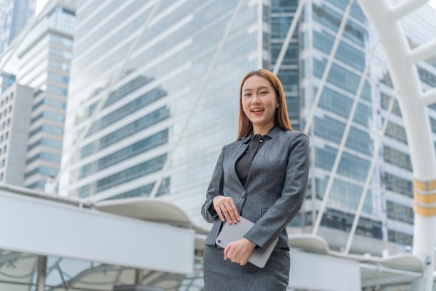 一名年轻的亚洲女商人穿着灰色的办公套装，在商业区使用她的平板电脑时微笑着图片下载