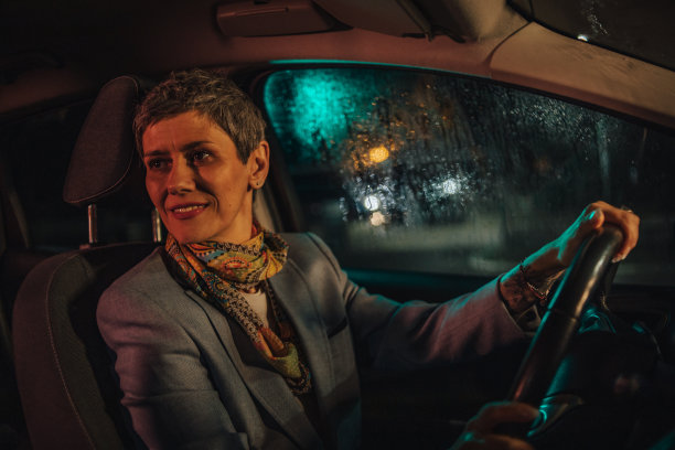 一个成熟的女人在晚上开车，城市的灯光和其他汽车在车窗上刺眼图片下载