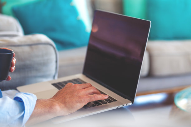 一个成熟的男人在家里的沙发上用笔记本电脑工作，喝着咖啡。图片下载