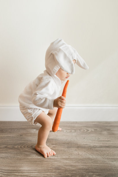 15个月大的古巴裔美国男婴，浅棕色短发，棕色眼睛和12只脚趾，穿着白色兔子毛衣，蓬松的兔子耳朵连接到头巾和短裤，赤脚坐在地板上放松，在自然光下嚼胡萝卜图片下载