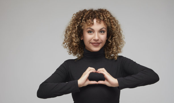 一头卷发的年轻女子高兴地用手做着心形手势。图片下载
