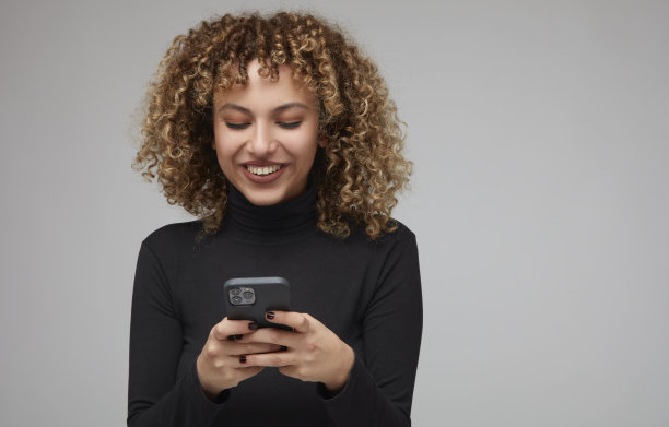 年轻的卷发女子高兴地看着她的手机上的东西。图片下载