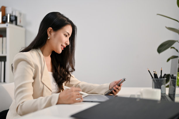 侧视图的亚洲女商人使用她的手机和平板电脑在她的办公桌。图片下载