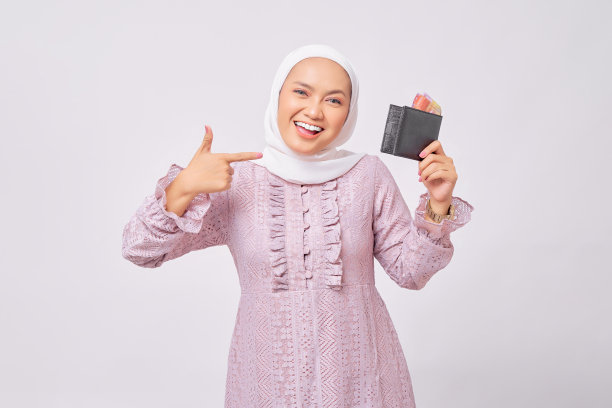微笑着美丽的年轻的亚洲穆斯林妇女戴着头巾和紫色连衣裙指着钱包的全部现金孤立在白色的工作室背景图片下载