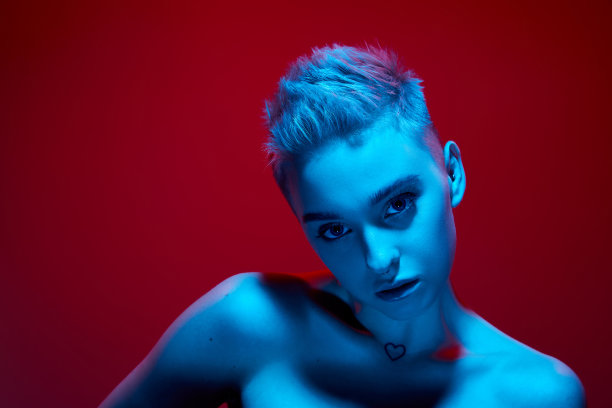 独特性。美丽的年轻女孩与短金发裸露的肩膀对红色工作室背景在蓝色霓虹灯。朋克风格图片下载
