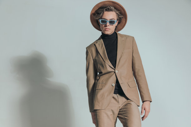 酷酷的年轻人在米色西装，戴着太阳镜和帽子，摆姿势图片下载