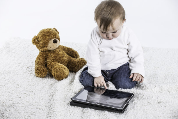 白色背景的工作室照片，一个婴儿触摸平板电脑的屏幕，旁边有一只泰迪熊图片下载