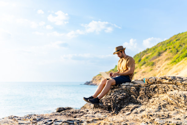 白人男子坐在海岸线山上，一边喝着热咖啡，一边在笔记本电脑上工作。图片下载