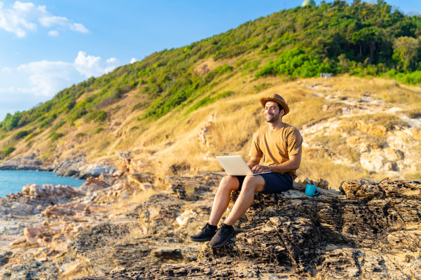 白人男子坐在海岸线山上，一边喝着热咖啡，一边在笔记本电脑上工作。图片下载