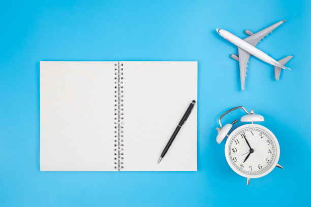 空白笔记本，闹钟和飞机在蓝色背景，平铺。图片下载