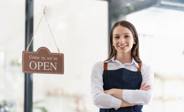 快乐的商业女人是一个围着围裙的女服务员，咖啡馆的老板站在门口，举着“开门”的牌子等待顾客。小型商业概念，咖啡馆和餐馆图片下载