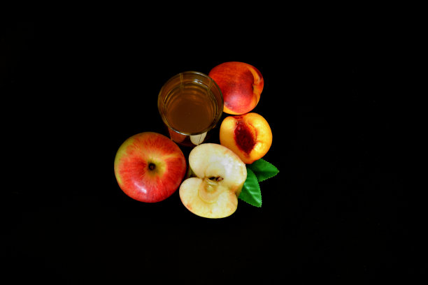 一杯黑色背景的果汁，旁边是一个红苹果和一个成熟的带叶子的桃子。图片下载
