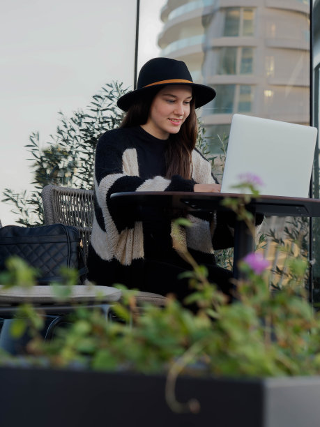 年轻的白人女孩在鲜花环绕的室外露台桌子上微笑着用笔记本电脑打字。电视工作。垂直的。副本的空间。图片下载