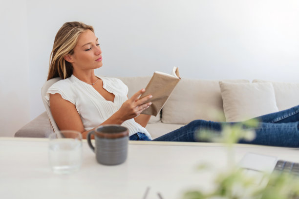 小女孩一边看书一边喝咖啡，把脚放在桌子上。桌子上放着书，背景是女人和笔记本电脑。图片下载