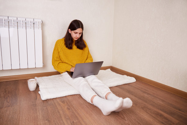 一个女孩坐在暖气片旁边的地板上，用笔记本电脑工作。图片下载