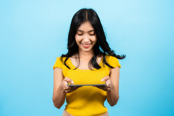 画像上，长发女孩穿着衬衫黄色紧身衣，手里拿着笔记本电脑，双手捧着，玩着有趣的游戏，蓝色背景上孤立的室内工作室。图片下载