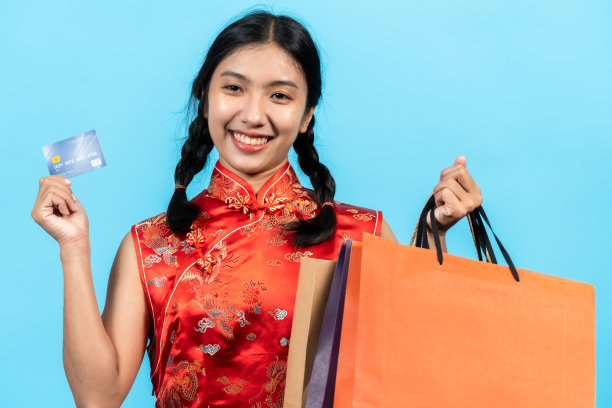 女孩在春节穿着旗袍，手里拿着信用卡和购物后的许多纸袋，他的脸看起来很高兴。室内工作室孤立的蓝色背景图片下载