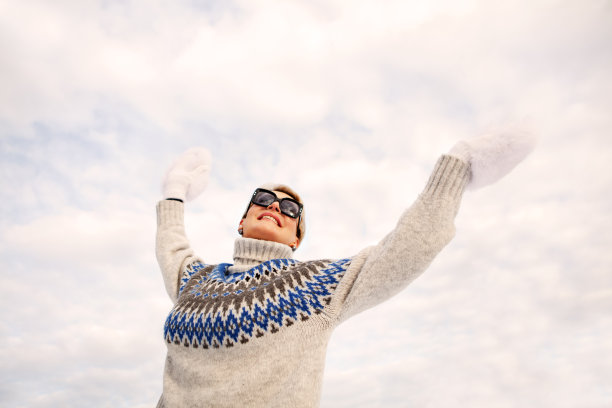 快乐的女人在冬装毛衣和眼镜举起手，跳舞和享受阳光对着白色多云的天空，积极的生活方式图片下载