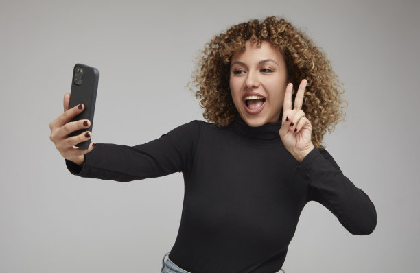 年轻的卷发女子做和平手势和鬼脸，同时用手机自拍。图片下载