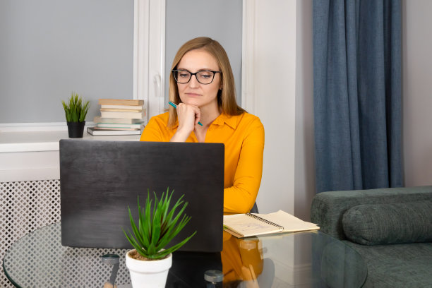 一位年轻专注的女士，戴着眼镜，穿着橙色衬衫，坐在一张玻璃桌子前，用笔记本电脑工作，手里拿着一个记事本记笔记。商业概念，心理学，市场营销，培训，发展。图片下载
