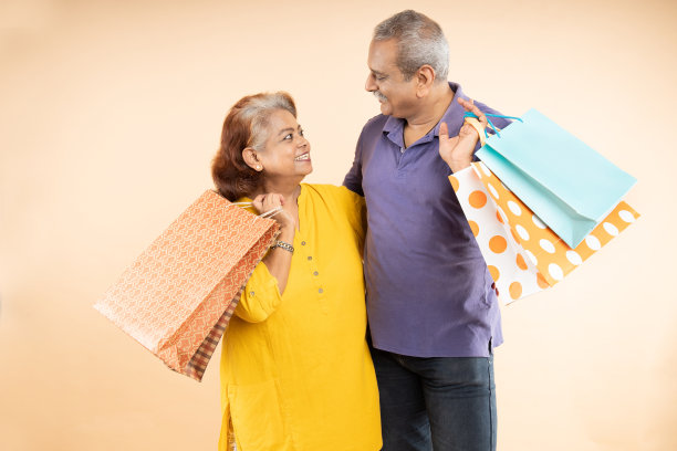 快乐的印度老夫妇拿着购物袋看着彼此孤立的米色背景。退休生活，电子商务销售和优惠图片下载