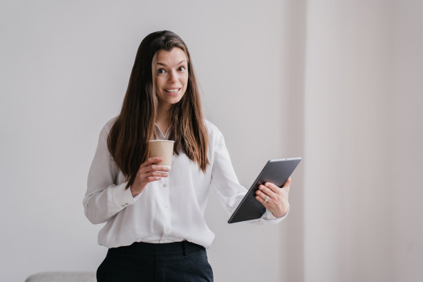 年轻的棕发女商人，穿着白衬衫和黑裤子，手里拿着平板电脑和一杯咖啡，在办公室靠墙看着相机露出牙齿的微笑。聪明的白人女学生被隔离。图片下载