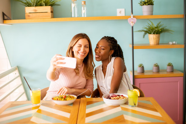 两名年轻女子，白人和黑人，在咖啡馆里用手机自拍图片下载