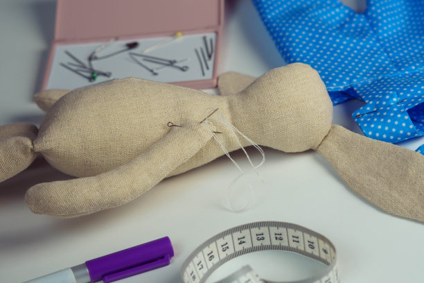 缝纫创造的填充兔子玩具与针在身体上躺在桌子上。针织的爱好。兔子玩具附近的蓝色波点纺织品图片下载