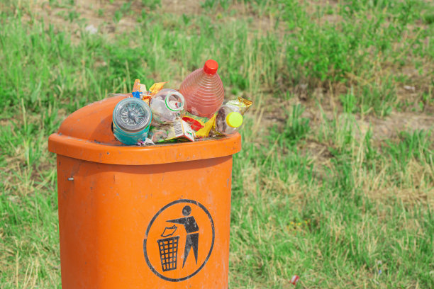 橙色的垃圾桶里堆满了垃圾瓶和废纸，背景是绿色的草地，等待着被送去回收和处理图片下载