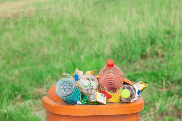 橙色的垃圾桶里堆满了垃圾瓶和废纸，背景是绿色的草地，等待着被送去回收和处理图片下载