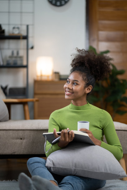 年轻的非裔美国妇女在家里的沙发上喝茶看书。图片下载
