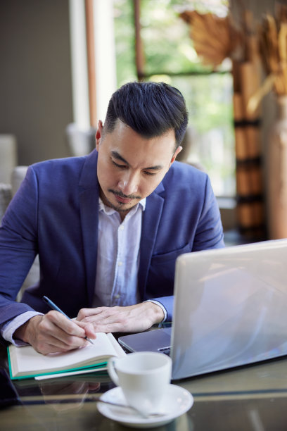 年轻英俊的亚洲商人使用笔记本电脑和写笔记，坐在咖啡馆或联合办公空间。电话会议或在线工作图片下载