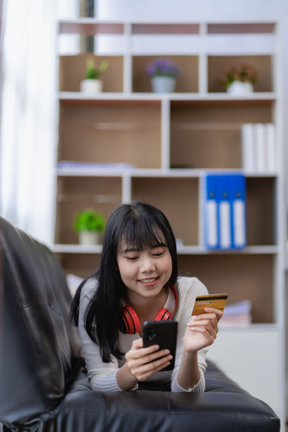 亚洲年轻女子手持信用卡支付电子货币远程网上购物在家里的沙发垂直图像图片下载