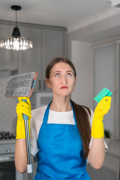 穿着制服的年轻女子满怀希望地抬起头，手里拿着拖把和海绵。不愿意打扫公寓，打扫后疲劳。垂直图图片下载