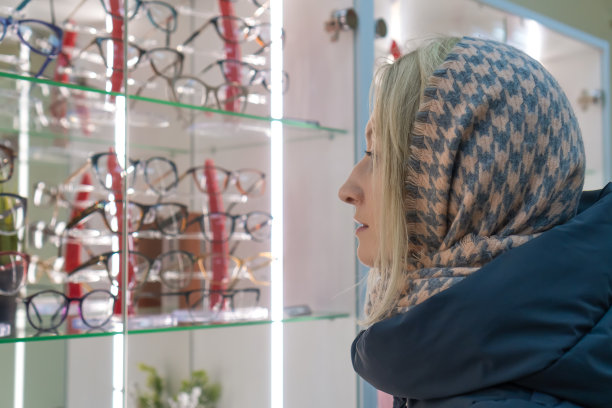 一名身穿夹克、头戴围巾的年轻金发女子在一家眼镜店为自己挑选眼镜。图片下载