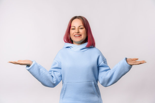 迷人的微笑的女人显示平衡的手。可爱的女模特，粉红色的头发，蓝色的卫衣，站在白色的背景图片下载
