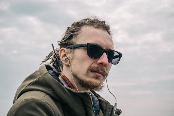 肖像快乐的人戴着眼镜和脏辫入耳式耳机或耳机听音乐。年轻的潮人看着相机图片下载