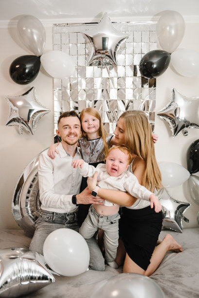 快乐的家庭庆祝生日聚会。爸爸妈妈祝贺女儿、儿子生日近拱。母亲和父亲拥抱着孩子。照片墙面装饰白色、黑色、银色气球。婴儿哭了。图片下载