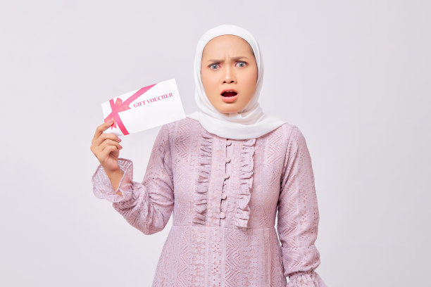 惊讶美丽的年轻的亚洲穆斯林妇女戴着头巾和紫色连衣裙拿着礼券证书，看着相机孤立在白色的工作室背景图片下载