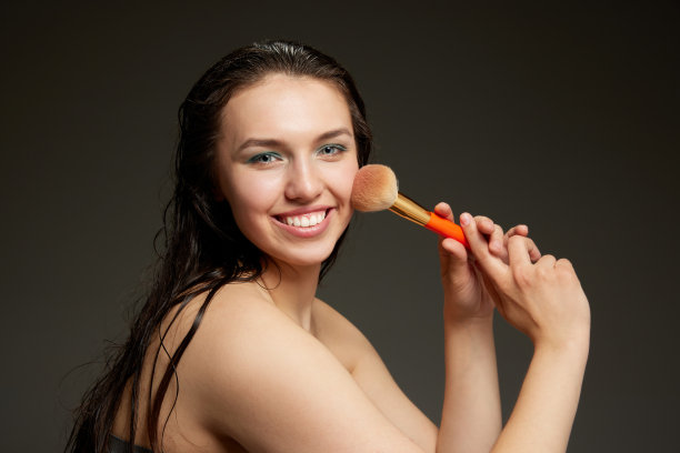 半长的肖像，年轻的情感美丽的女孩拿着腮红刷，做化妆孤立在灰色的背景。美丽，时尚，风格，乐趣和快乐的概念图片下载