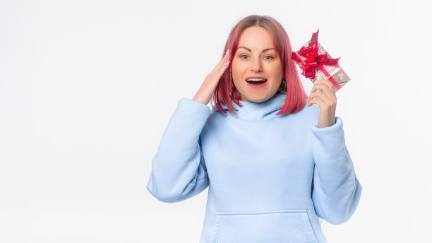 快乐的年轻女子，粉红色的头发，穿着蓝色的连帽衫，抱着摇礼盒与礼品丝带蝴蝶结试图猜什么里面，站在白色的背景图片下载