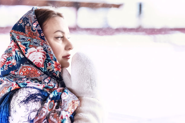 一个斯拉夫妇女围着民族色彩的围巾，披着毛皮大衣，戴着温暖的手套，站在一座木屋的门廊上。冬天,下雪图片下载