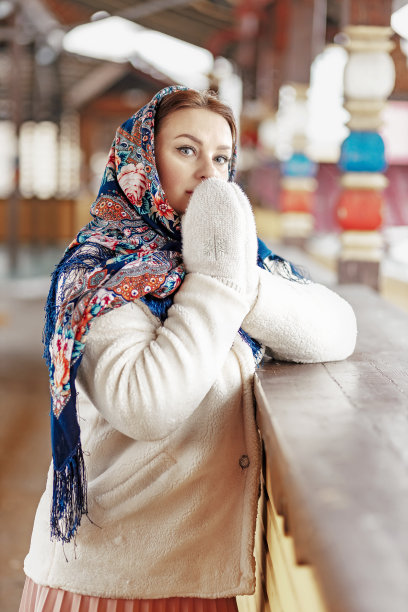 一个斯拉夫妇女围着民族色彩的围巾，披着毛皮大衣，戴着温暖的手套，站在一座木屋的门廊上。冬天,下雪图片下载