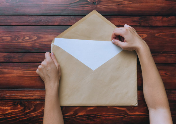 两个女人的手拿着一个大的牛皮纸信封，里面有一张空白的白纸，放在棕色的木制背景上。空白字母概念与复制空间。图片下载