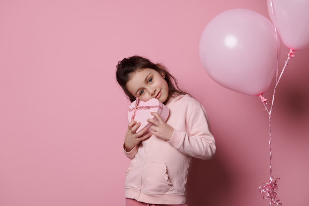 美丽的小女孩，享受节日活动，拿着一个心形的礼品盒，在粉红色的背景下微笑图片下载